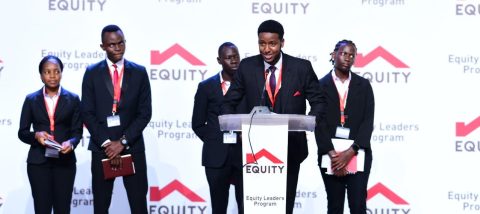 A man in a suit speaks into a microphone from a podium. Standing behind him are men and women also in suits. The background reads "Equity Leaders Program".