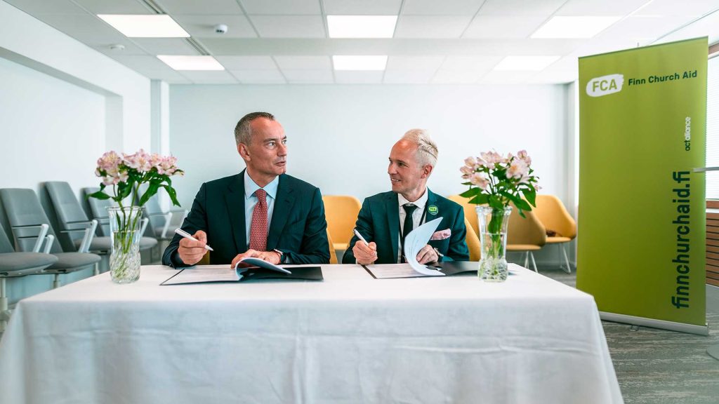 Two men sit behind a table signing a paper. There are flower vases on the table and chairs in the background.