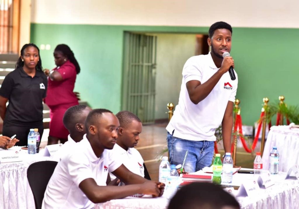 A man in a white polo shirt bearing the logo "Equity" stands at a table in a conference room and talks into a microphone.