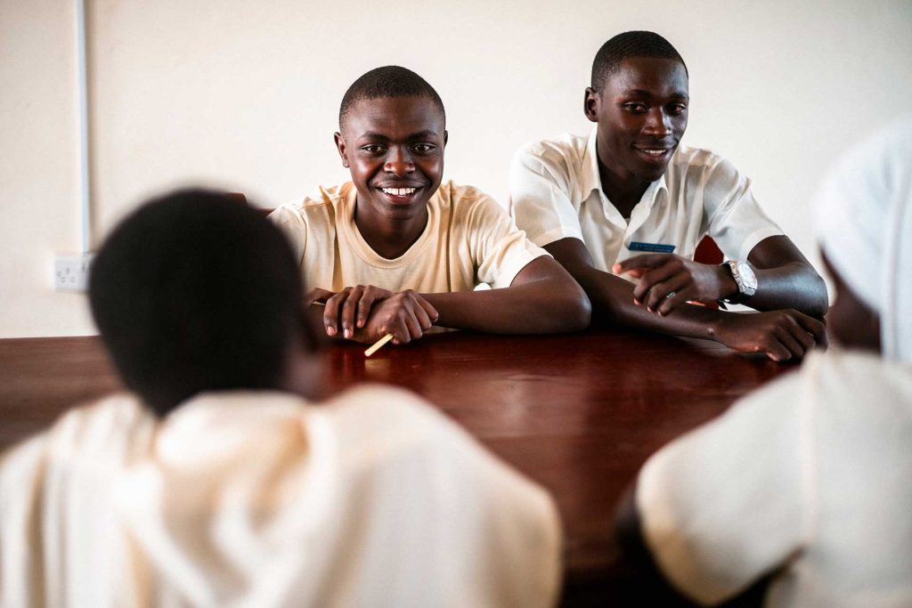 Kaksi poikaa hymyilee luokkahuoneessa pulpetin takaa edessään istuville tytöille, jotka on kuvattu takaapäin. 