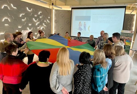 A number of people in a meeting room wearing lanyards are holding a rainbow-coloured parachute.