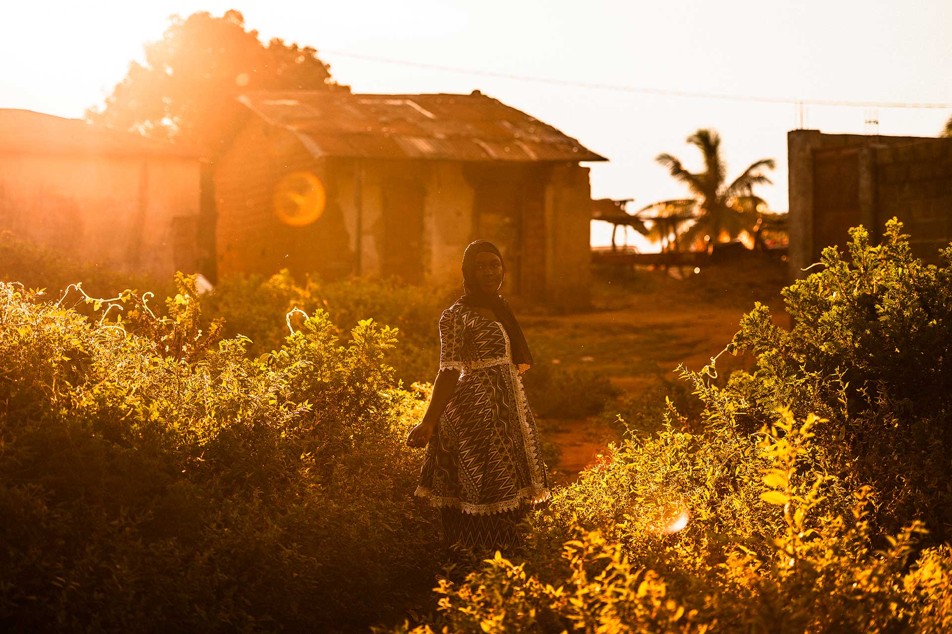 Nuori nainen seisoo auringonlaskun valaisemalla pihalla Keski-Afrikan tasavallassa. Taustalla näkyy rakennus. A young woman stands in a courtyard lit by sunset in the Central African Republic, with a building in the background.