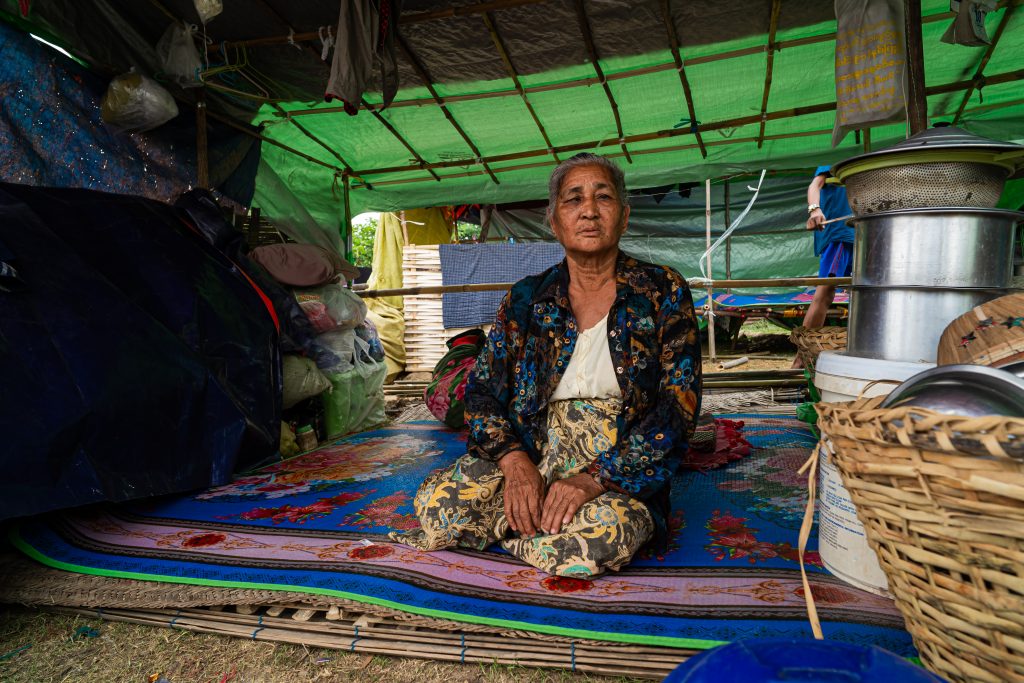An elderly Burmese lady sits on a carpet in a temporary shelter