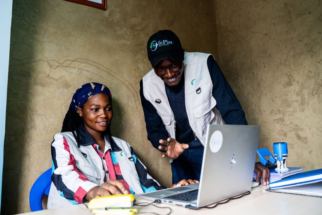 A woman sitting at a computer working, and a man standing next to her pointing and smiling.