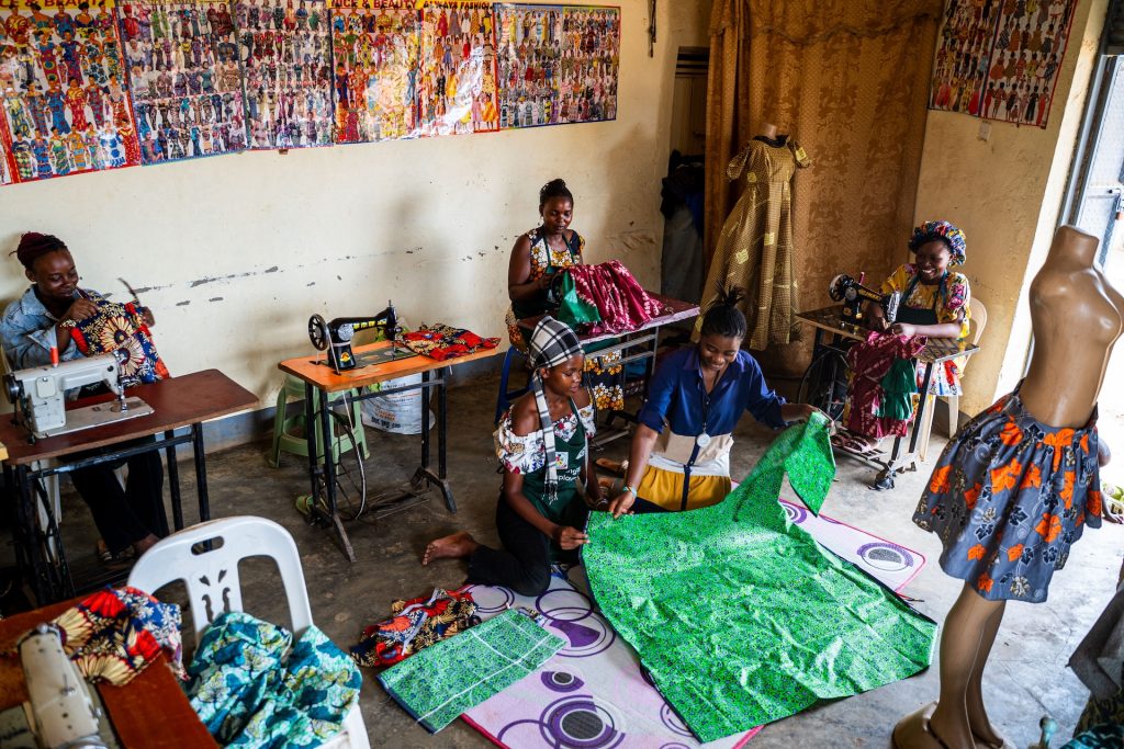 Three women sit at their sewing machines while two women sit on the floor working with cloth.