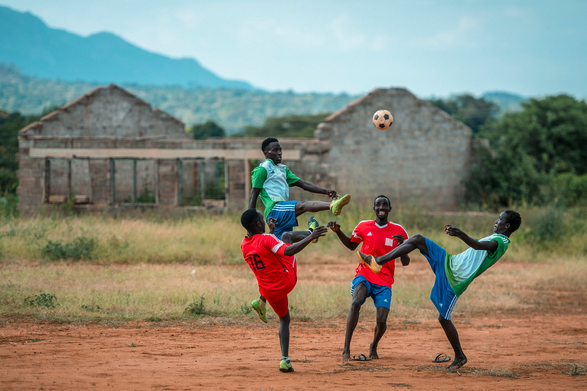 Thre men playing football on a gravel pitch with some mountains and buildings in the background.