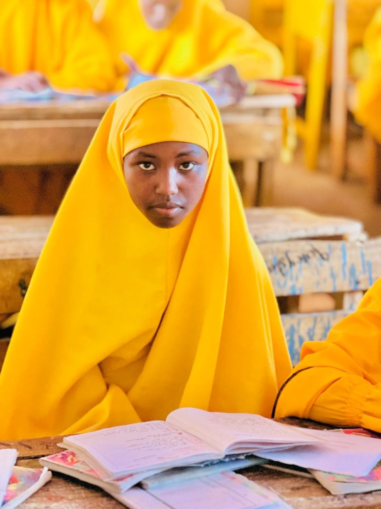 A schoolgirl in a yellow hijab and abaya sits at a classroom desk.