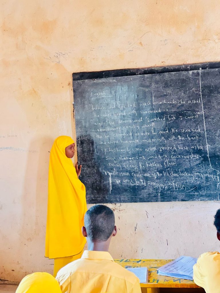 A girl in a yellow hijab and abaya stands next to a blackboard.