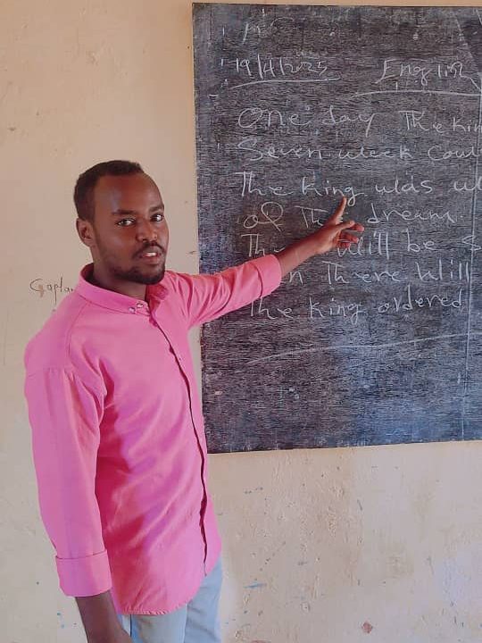 A man in a pink shirt gestures at a blackboard