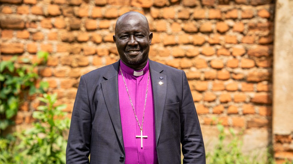 A man in a Bishops's outfit with a large cross around his neck standing in front of a tile wall
