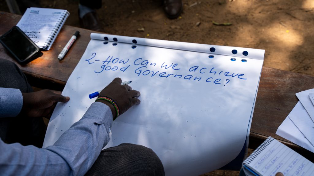 A hand writing on a large sheet of paper. The text on the paper says "2. How can we achieve good governance?"