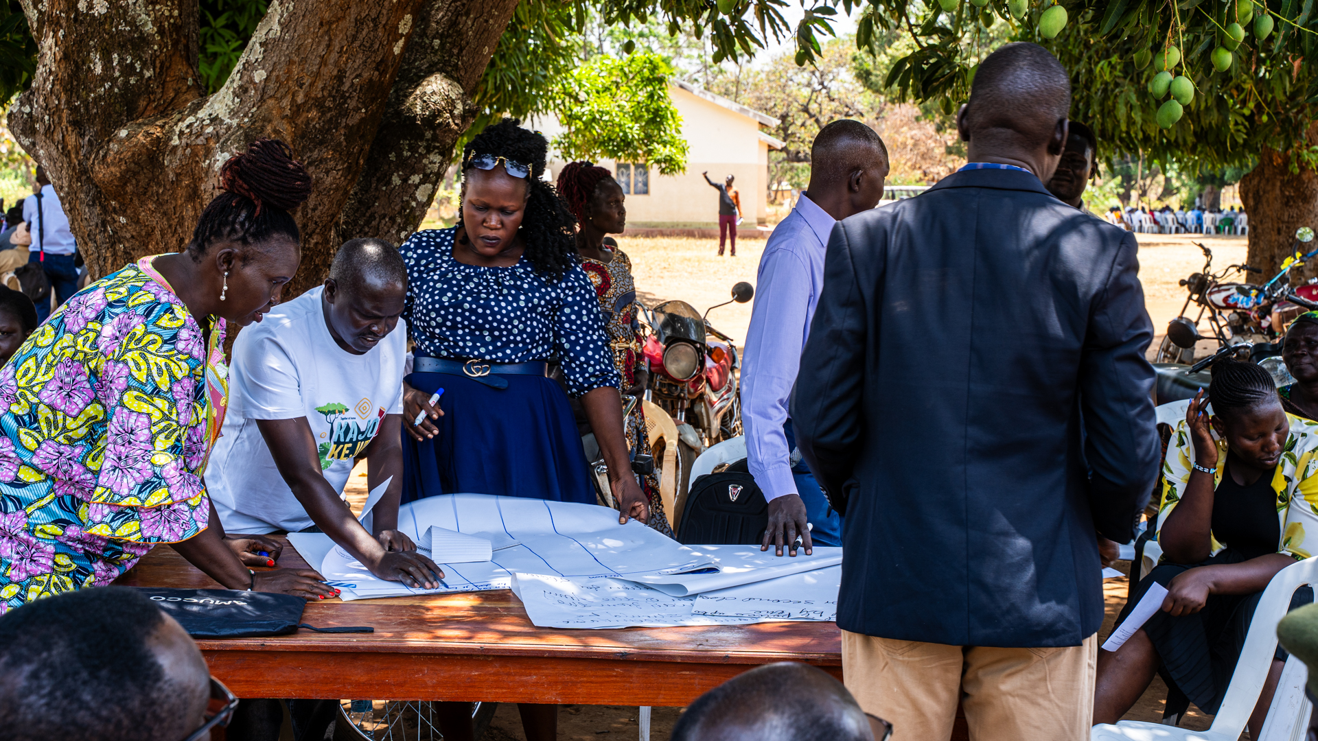 A man writing on a big sheet of paper with two women looking over his shoulder. MAny people are standing around or sitting on plastic chairs.