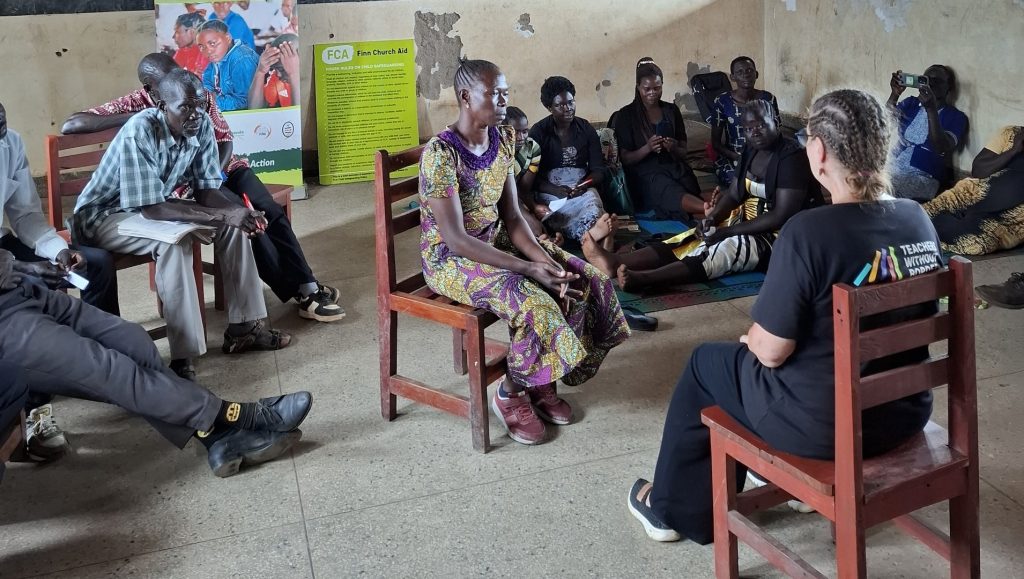 A number of people sit in a bare room on chairs or on the floor.