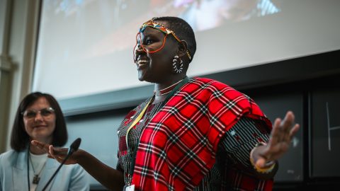 A woman in Kenyan national dress gestures and smiles while giving a talk.