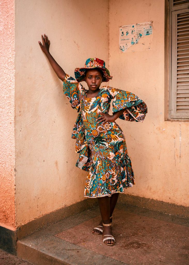 A woman in a patterned dress with frills and ruffles and a matching hat leans against a wall.