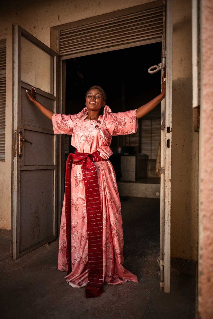 A woman in a pink gown with a red belt stands with arms outstretched in a doorway.