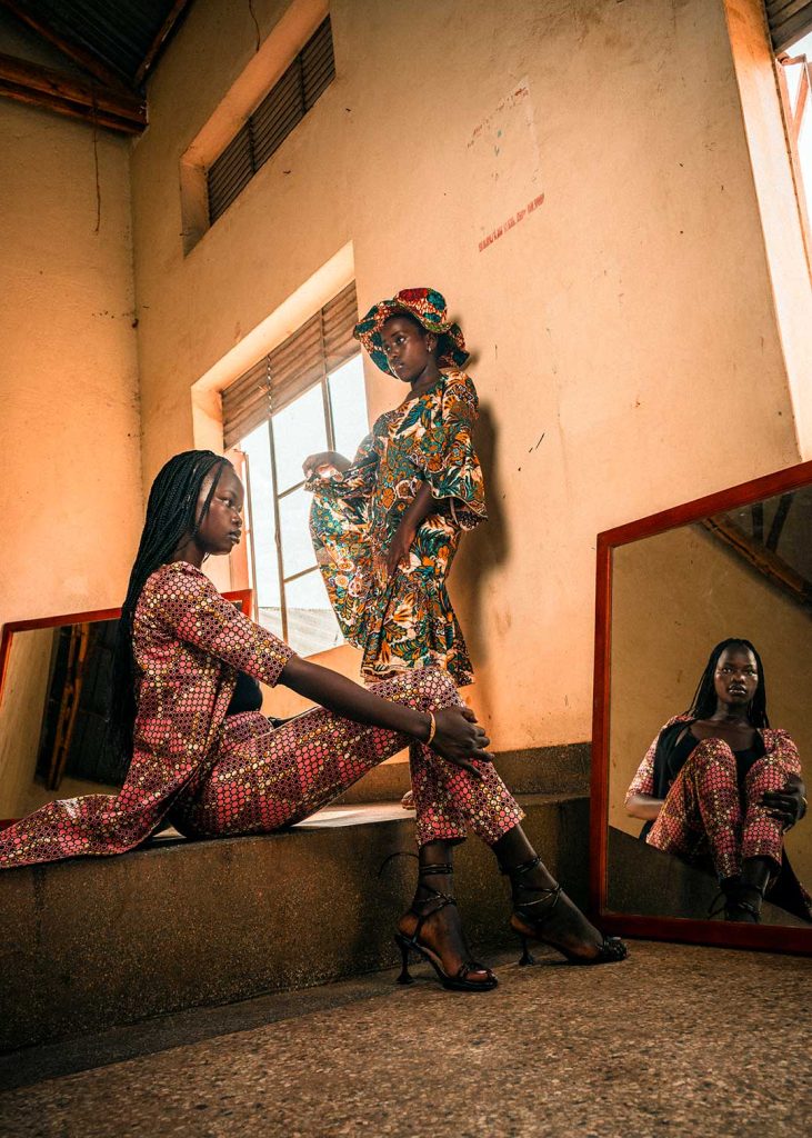Two women pose for a fashion photo. The woman in the foreground is sitting on a podium, the woman in the background is leaning against the wall. The image of the seated woman is reflected in a mirror on the floor.