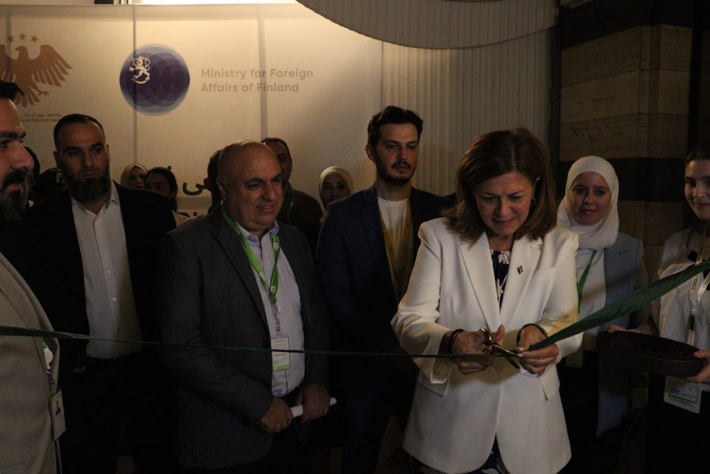 A man and a woman stand in a room in front of a taut ribbon, which the woman is cutting