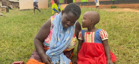 A girl sits on a lawn with an adult woman, whispering in her ear.