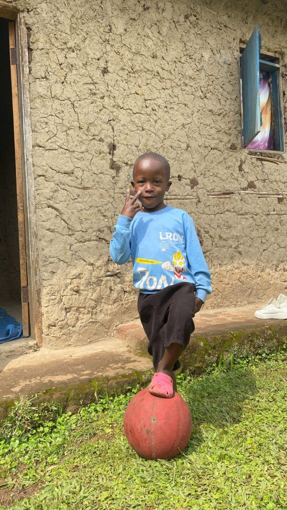 A young boy standing next to a house on a lawn with a football under one of his feet.