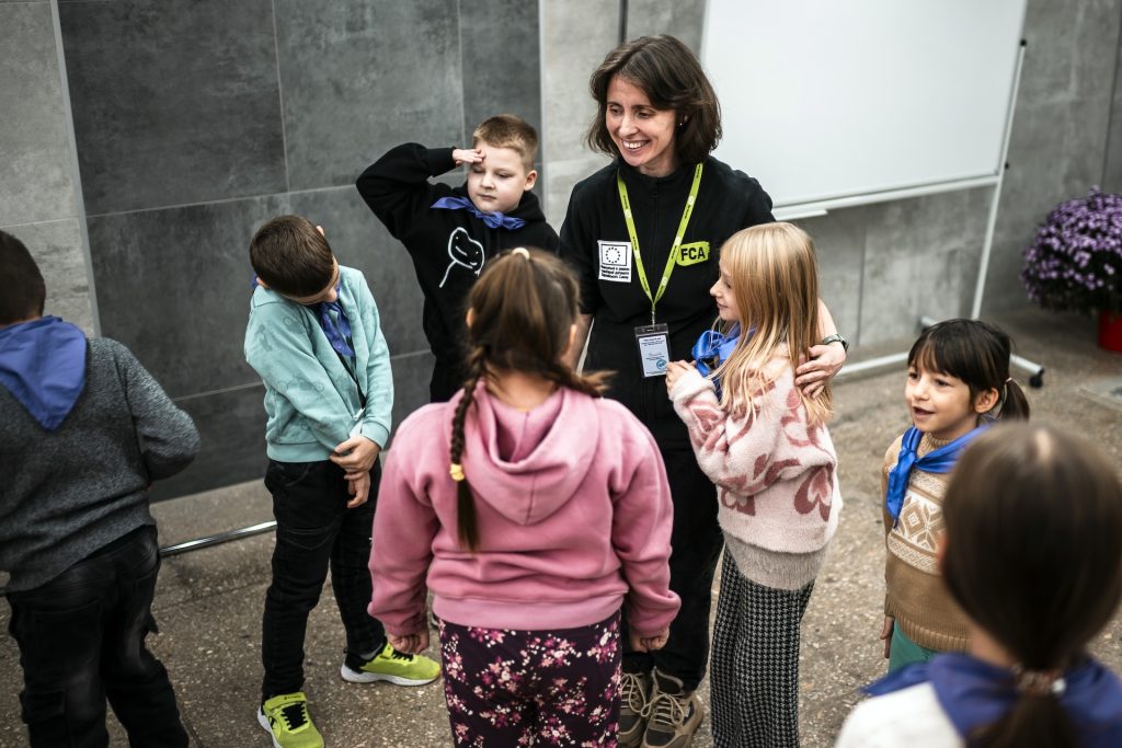 A smiling woman wearing an FCA branded shirt stands in the middle of a group of children.