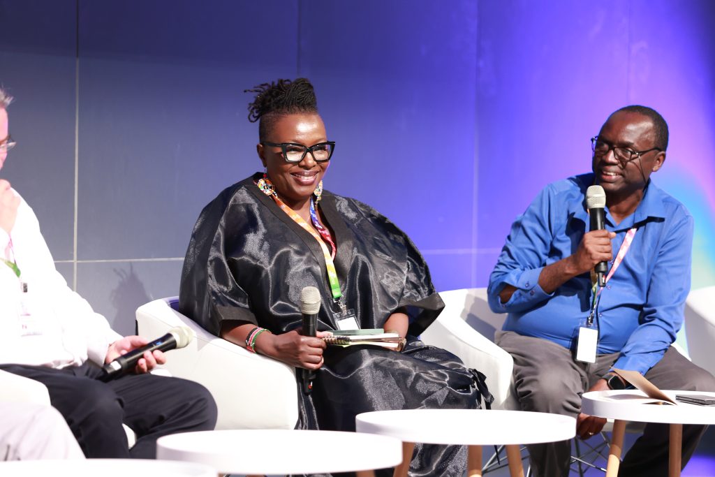 A woman and a man sit on chairs behind small tables. Both hold microphones and are smiling. The man is holding the microphone to his mouth as if about to speak.