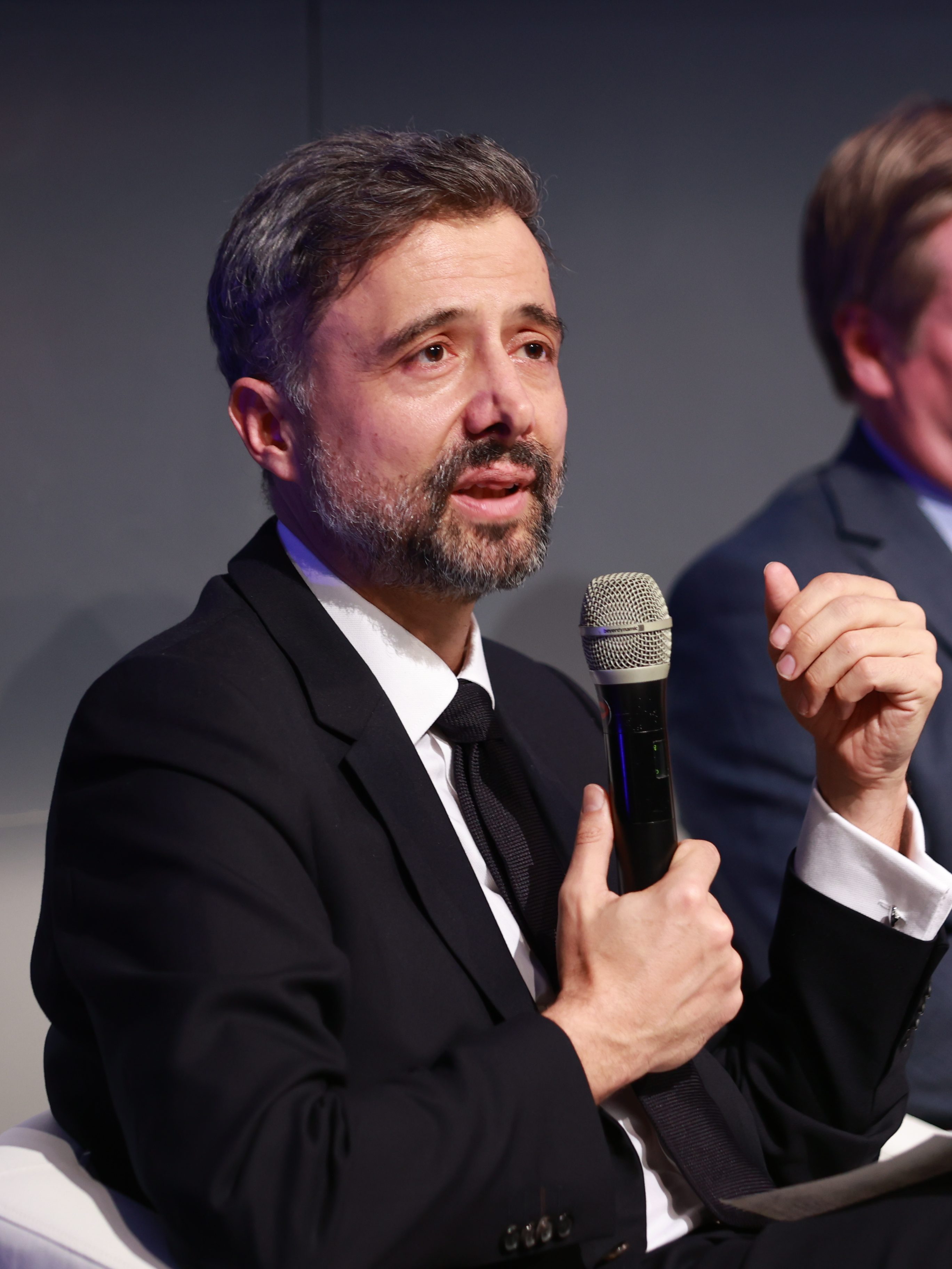 A man in a business suit sits while speaking into a microphone. A woman and a man are listening in the fore- and background.