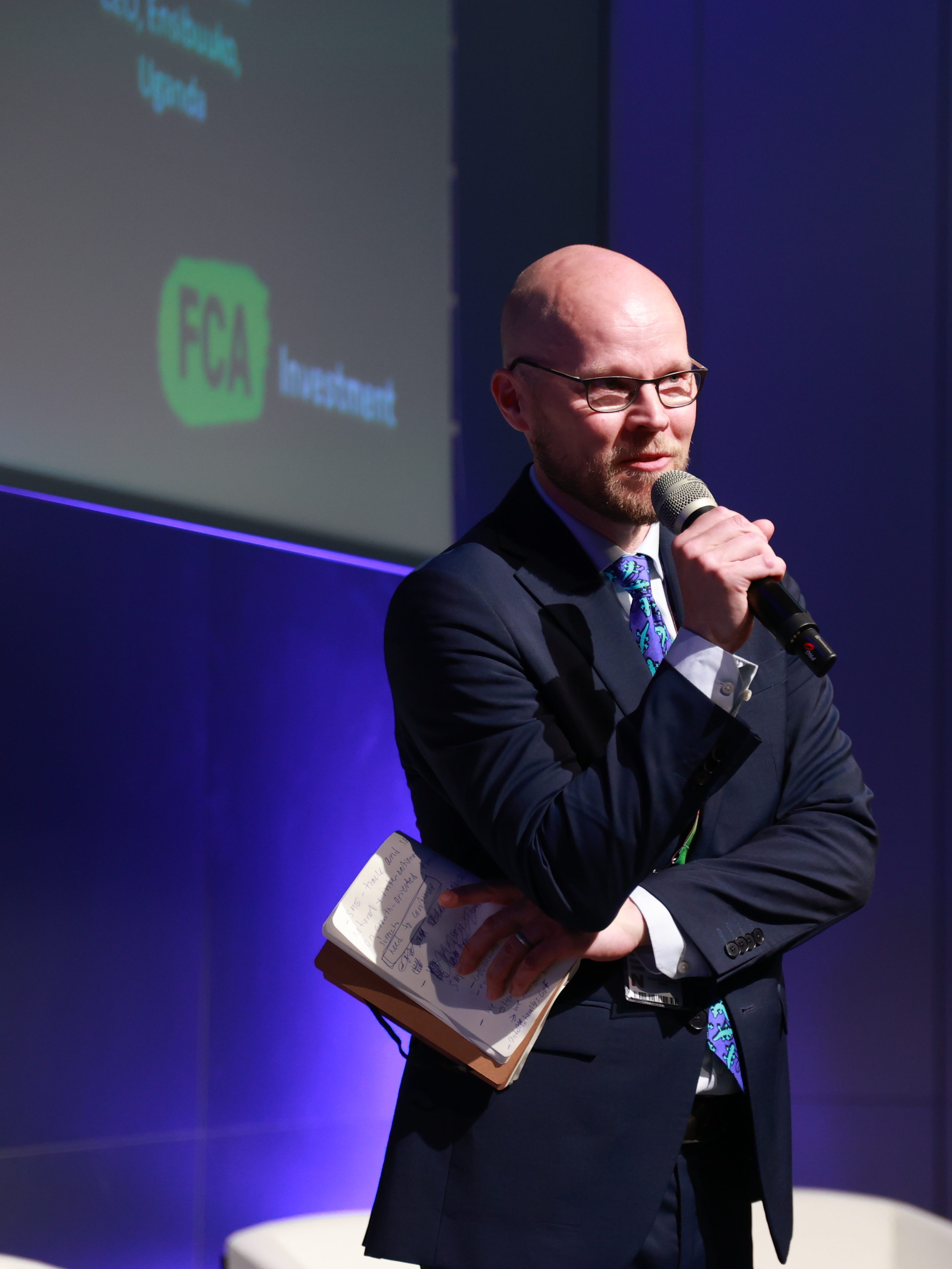 A man with a microphone speaks on a stage. Behind him we see the logo of FCA Investments