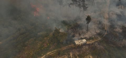 A picture from above of a fire truck driving in a partially burned down forest. A part of the forest is still on fire.