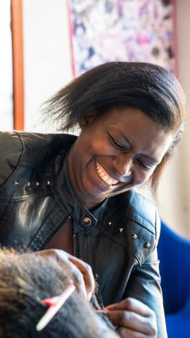 A smiling woman in a salon putting a braid in another woman's hair.
