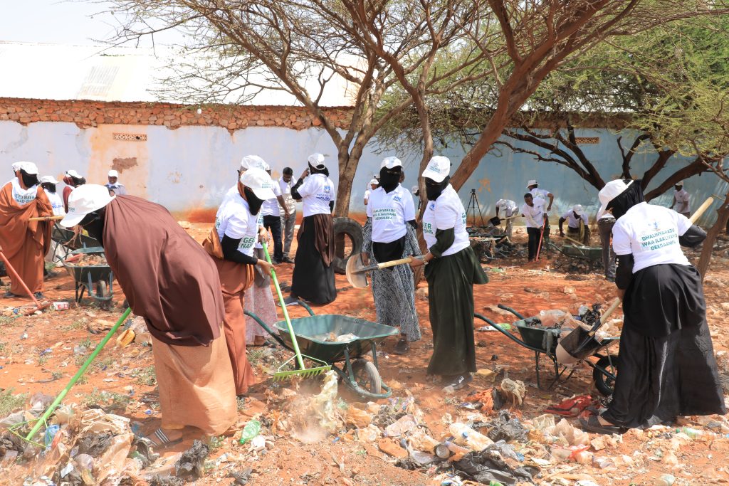 A group of people, mostly female, cleaning a site next to a concrete wall and collecting trash