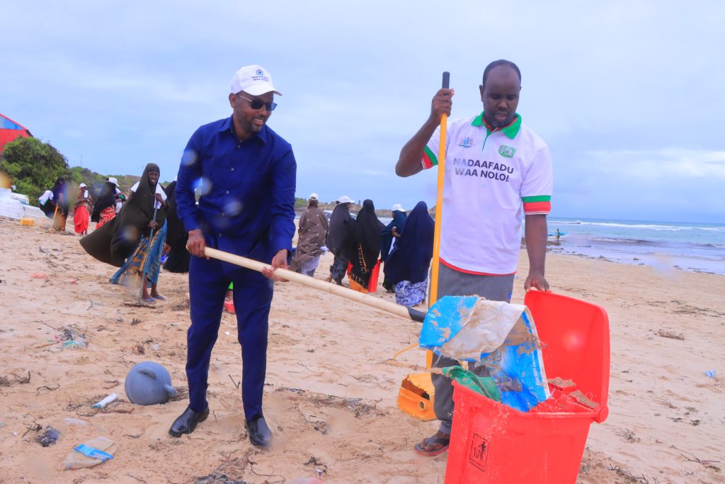 Two men collecting trash on a beach next to the water. Behind the two men, a large group of women are also participating in the cleaning.