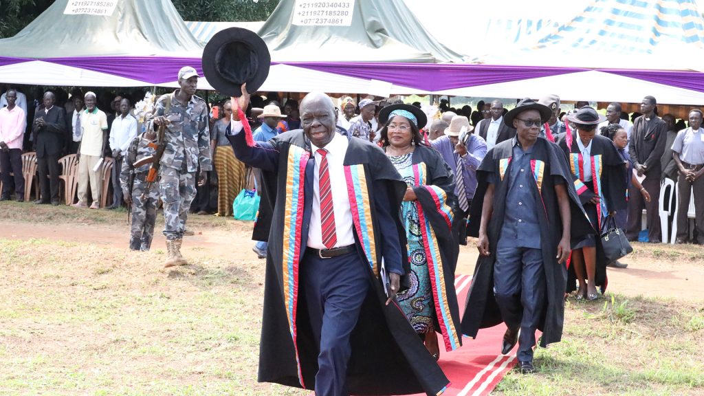 A man in a suit with his hat lifted in his hand above his head walks the red carpet at a graduation ceremony.
