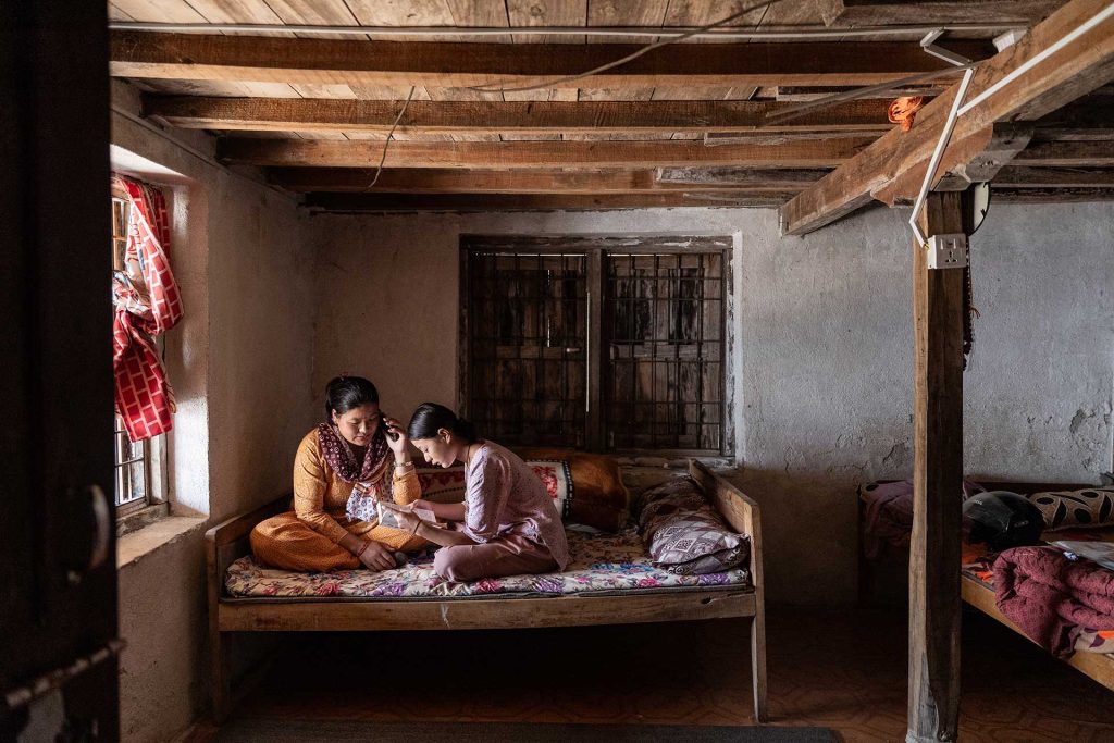 Nepalilaiset nainen ja tyttö istuvat sängyllä. Tyttö lukee ja äiti kuuntelee vieressä.