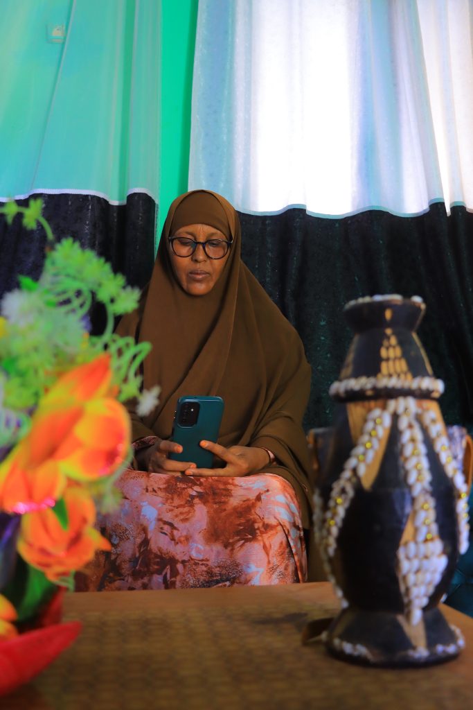 A woman in an abaya and hijab sits on a sofa looking at her phone. In front of her is a low coffee table containing some ornaments.