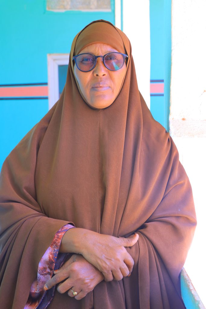 A woman in a brown abaya and hijab looks seriously at the camera