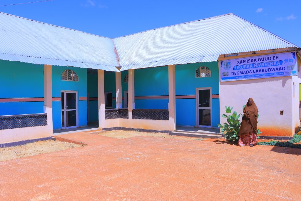 A woman stands next to a long blue building in the bright sunshine.