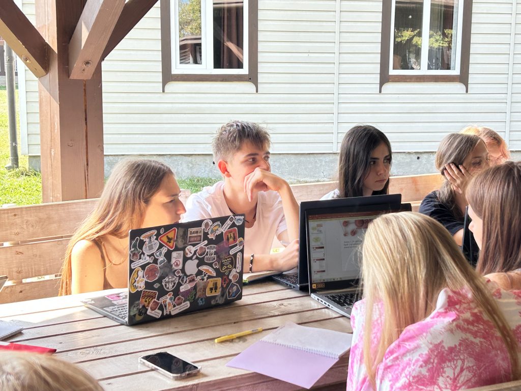 A group of teenagers sit at outside tables, in front of laptops, listening to someone speaking off-camera.