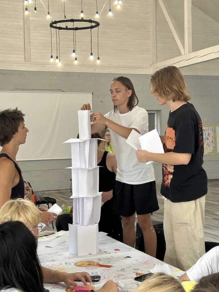 Teenagers look on as a boy builds a tower out of paper on a desk.