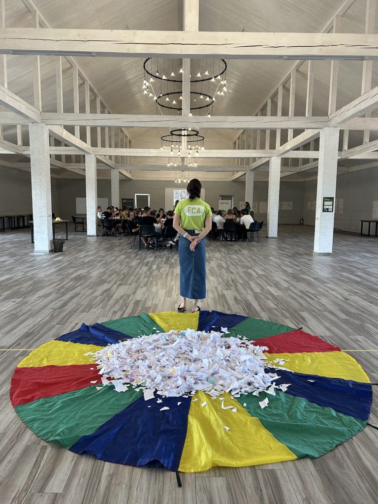 In the foreground is a rainbow parachute lying on the ground. In the middle of the parachute are a large number of scraps of paper. In the middleground a person in an FCA branded shirt stands looking on at a group of students sitting on chairs in a large hall. 