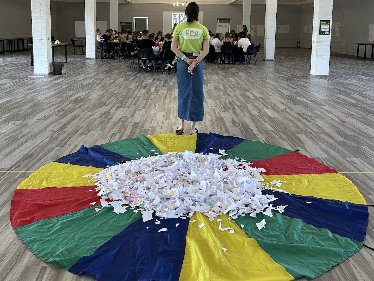 In the foreground is a rainbow parachute lying on the ground. In the middle of the parachute are a large number of scraps of paper. In the middleground a person in an FCA branded shirt stands looking on at a group of students sitting on chairs in a large hall.