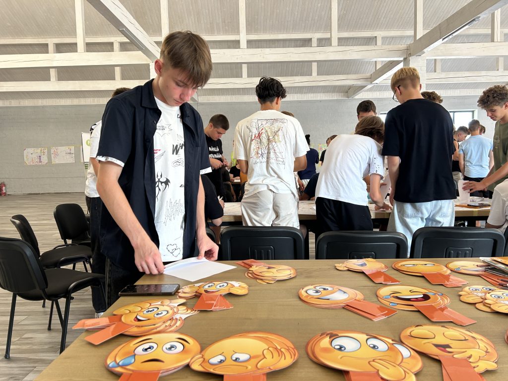 A teenaged boy stands at a table full of printed out emojis. In the background, other teenagers busy themselves arranging papers.