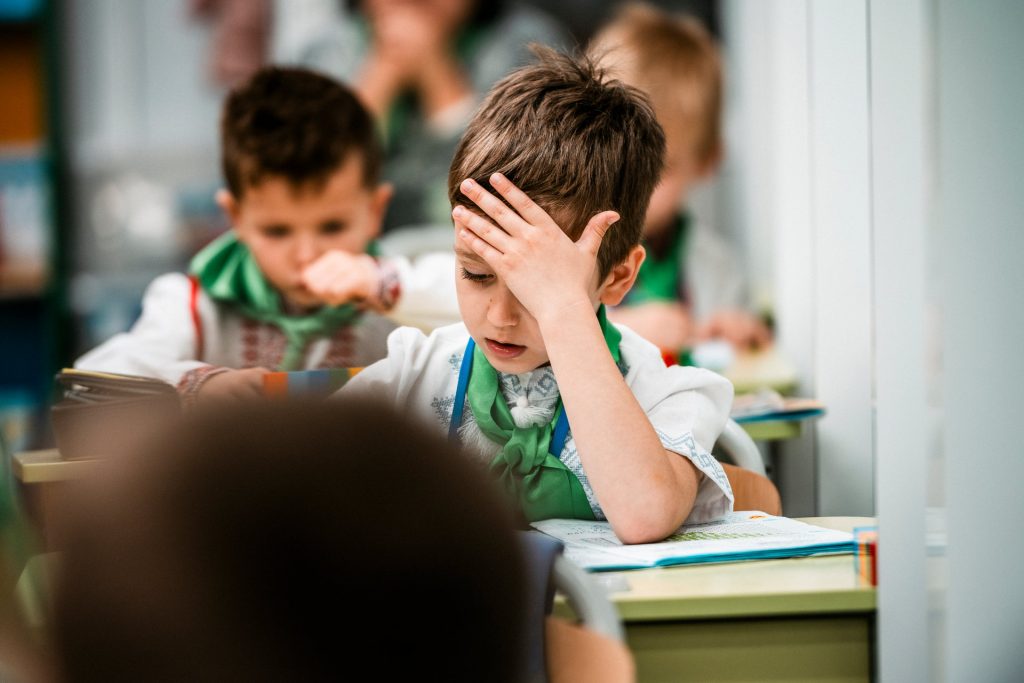 A schoolboy sits at a desk and puts his hand over his head as he concentrates on the work on his desk.