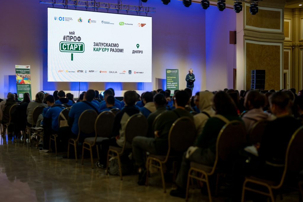 Many people sit in an auditorium looking at a stage. Behind the stage is a large screen with the ProfStart logo and many smaller logos of supporting organisations.