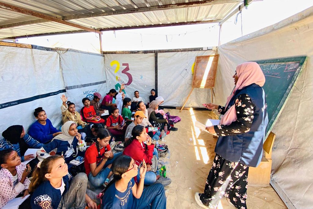 Opettaja seisoo lattialla istuvien lasten edessä puhumassa. Luokkahuone on rakennettu suureen telttaan Gazassa. 