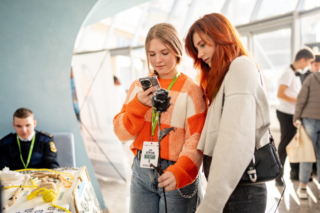 Two young women look at a mobile phone. They are wearing FCA lanyards and ProfStart ID cards.