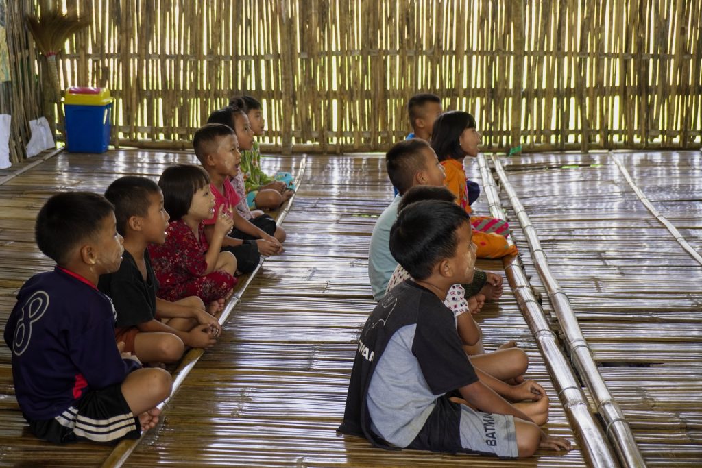 Children sit on a bamboo floor in rows in a classroom.