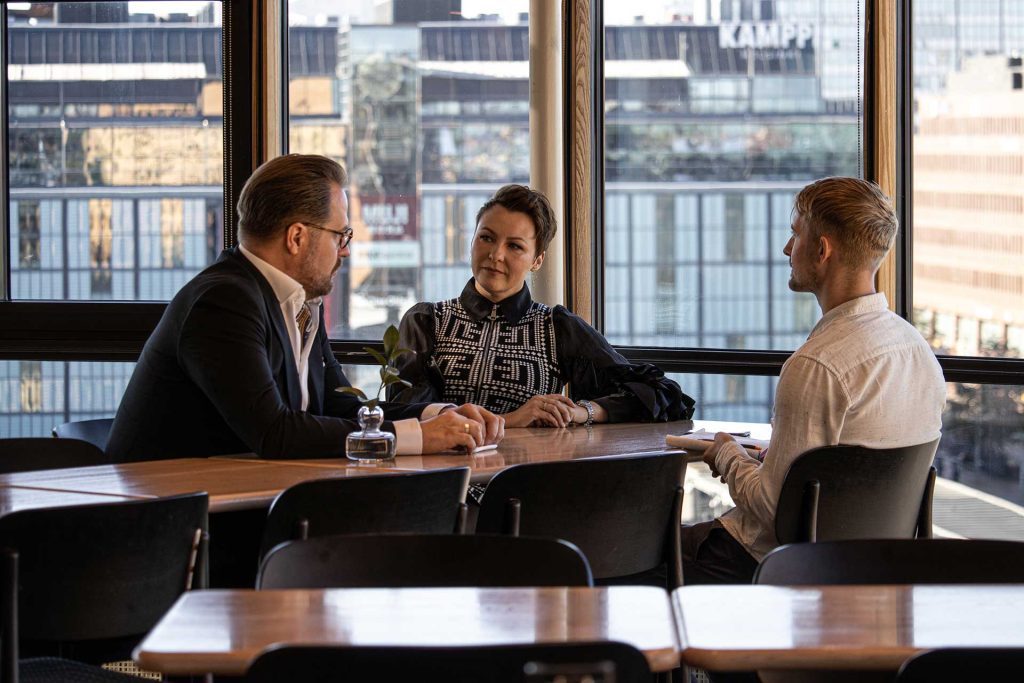 Kolme henkilöä istuu keskustelemassa pöydän ääressä. Taustalla olevista isoista ikkunoista näkyy ulos.