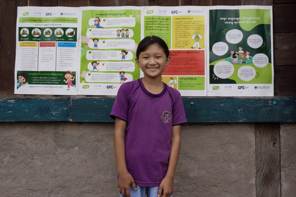 A smiling girl stands in front of a board with posters showing educational material for children bearing FCA and GPE's logos amongst others.