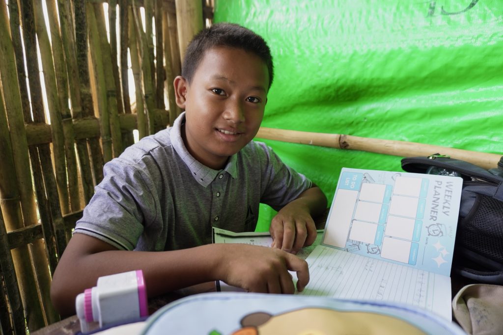 A smiling boy looks up from a textbook he is reading.